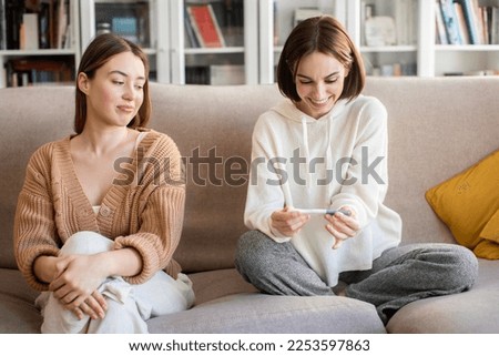 Smiling displeased european millennial lady looking at happy surprised woman with pregnancy test, rejoice positive result in living room interior. Envy, good news and baby waiting together at home Royalty-Free Stock Photo #2253597863