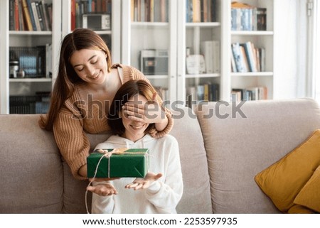 Cheerful european millennial lady closes eyes to her friend and gives gift box in living room interior, copy space. Celebration holiday, anniversary, birthday surprise and congratulations at home