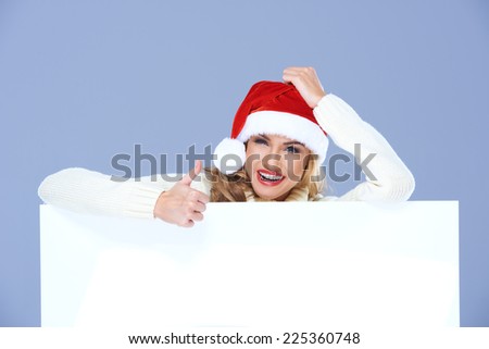 Happy Young Woman in Santa Hat Behind White Big Board  Giving Thumbs Up Gesture. Isolated on Light Blue Violet Background.