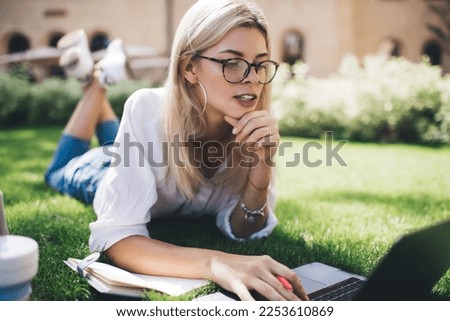 Millennial female student in optical spectacles using laptop application for making online research during e learning in campus park, skilled freelancer working remotely on digital netbook technology
