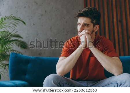Young minded sad man wear red t-shirt prop up chin look aside sit on blue sofa couch stay at home hotel flat rest relax spend free spare time in living room indoors grey wall. People lounge concept Royalty-Free Stock Photo #2253612363