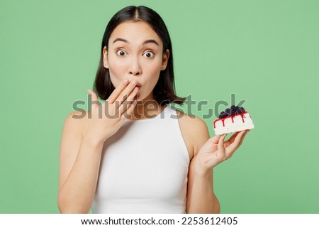 Young happy fun woman wear white clothes holding in hand pice of cake dessert cover mouth isolated on plain pastel light green background. Proper nutrition healthy fast food unhealthy choice concept
