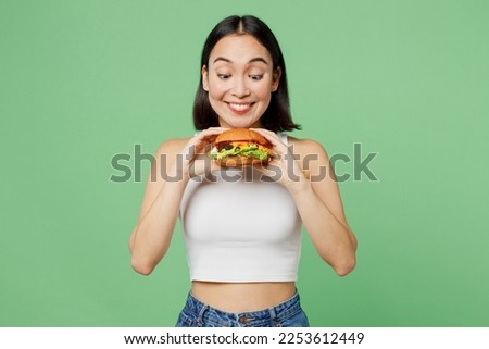 Young smiling happy excited woman wearing white clothes holding eating biting tasty burger isolated on plain pastel light green background. Proper nutrition healthy fast food unhealthy choice concept