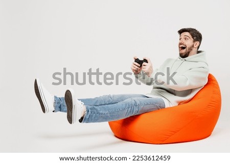 Full body gambling amazed young man wear mint hoody sit in bag chair hold in hand play pc game with joystick console isolated on plain solid white background studio portrait. People lifestyle concept