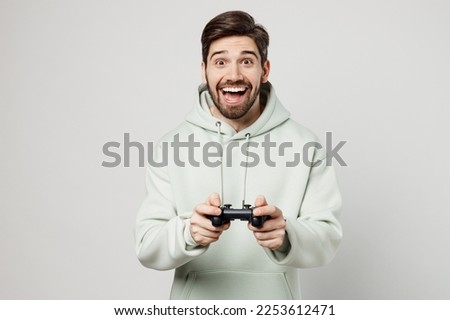 Young exultant excited fun caucasian man wear mint hoody look camera hold in hand play pc game with joystick console isolated on plain solid white background studio portrait. People lifestyle concept