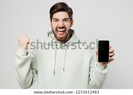 Young happy man wear mint hoody hold in hand use mobile cell phone with blank screen workspace area do winner gesture isolated on plain solid white background studio portrait. People lifestyle concept
