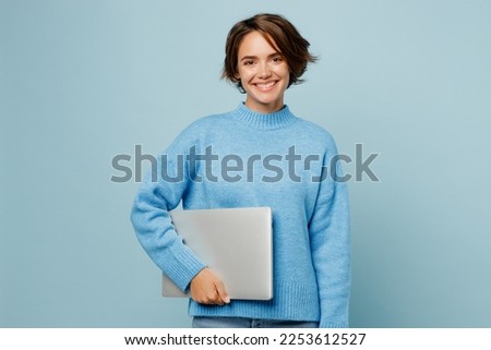 Young happy fun caucasian smart IT woman wear knitted sweater hold closed laptop pc computer look camera isolated on plain pastel light blue cyan background studio portrait. People lifestyle concept