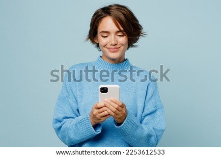 Young smiling happy fun caucasian woman wear knitted sweater hold in hand use mobile cell phone chatting isolated on plain pastel light blue cyan background studio portrait. People lifestyle concept Royalty-Free Stock Photo #2253612533