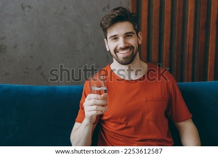 Young man wears red t-shirt hold drink clear fresh pure still water from transparent glass sit on blue sofa stay at home hotel flat rest relax spend free spare time in living room indoors grey wall