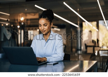 Positive African businesswoman sitting by the desk, typing something on the laptop. Royalty-Free Stock Photo #2253641659