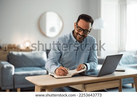 A cheerful male employee working over the laptop, writing notes in a notepad, sitting in the living room. Royalty-Free Stock Photo #2253643773