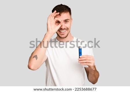 Young caucasian man holding a vaporizer cut out isolated excited keeping ok gesture on eye.