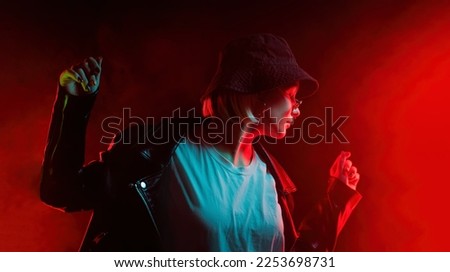 Fashionable woman dancing in red neon light, smoke background. Z generation enigmatic beautiful lady in leather jacket, panama hat, eyewear. High quality photo