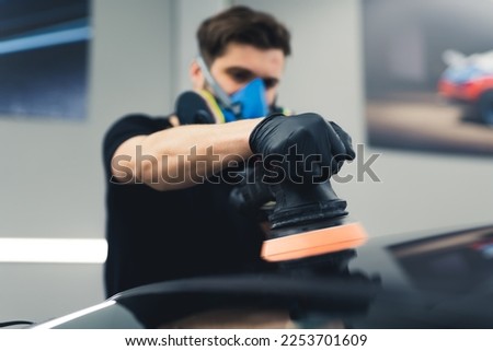 Caucasian man wearing protective face mask and black gloves using professional buffer tool to polish top of black car. Car detailing process. Horizontal indoor shot. High quality photo