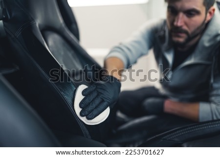 Caucasian man wearing black gloves leaning into car waterproofing leather car seat upholstery. Protective coating. Professional car detailing. Focus on foreground. Horizontal indoor shot. High quality