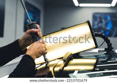 Unrecognisable man removing dents in car using professional equipment and lamp in a garage. Professional car repair. Car detailing. Horizontal indoor shot. High quality photo
