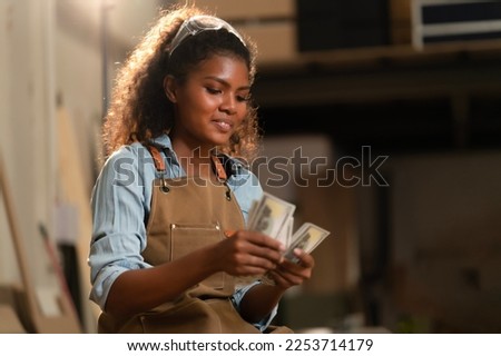 Young black carpenter woman counting money on hands after selling wood crafts handmade at her carpentry shop, receiving payment from customer by transferring her account. Selling wood furniture online