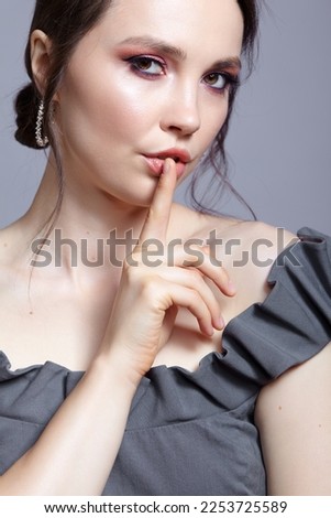 Portrait of brunette woman on gray background. Female in grey dress with pink smoky eyes eye shadows. Finger on lips with silence sign.