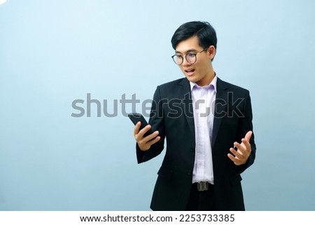Portrait of a surprised young smart asian businessman looking at his smart phone screen in disbelief with light blue background. Concept of business.