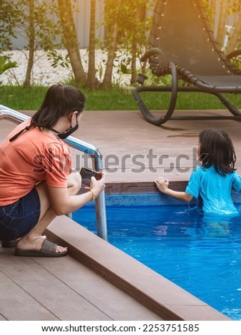Asian mother wear face mask use smart phone to take picture her lovely daughter in blue swimming suit play with clear blue water. Happiness mom and daughter at pool. Happy family relationship outdoor.