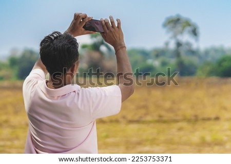Back view of Asian man using cell phone to take sky photo in field. Taking photo on mobile phone of sky view. Male take picture of the landscape with his smartphone on cloudy day. Outdoor happiness.