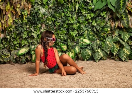 Asian little girl in red swimsuit in sunglasses sitting on sandy beach at tropical greenery, looking away. Young lady kid model in swimwear. Childhood summer vacation concept. Copy text space for ad