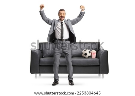Excited businessman cheering in front of a black sofa with a football isolated on white background