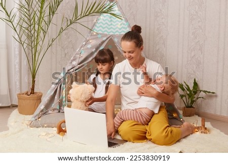 Horizontal shot of charming family, mother with her little daughters, sitting on floor near peetee tent and using laptop for watching cartoons. Happy childhood and motherhood.