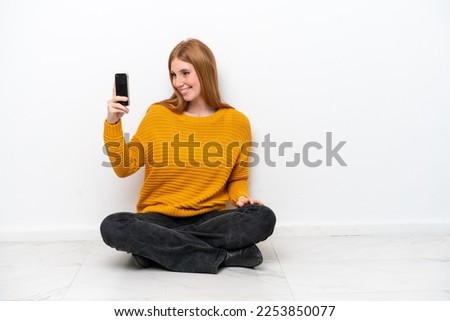 Young redhead woman sitting on the floor isolated on white background making a selfie