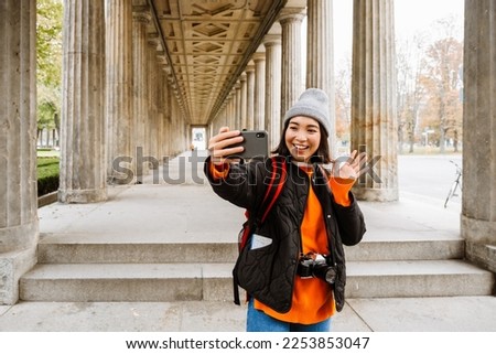 Excited young asian woman tourist with backpack taking selfie and waving during walk through the old city street
