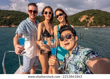 Group of diverse friend taking photo and drink champagne while yachting. Attractive young men and women hanging out, celebrating holiday vacation trip while catamaran boat sailing during summer sunset