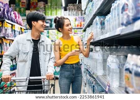 Asian young man and woman using cart to shoping goods in supermarket. Attractive couple picking product to purchase and walking together with happiness, enjoy buying in shopping mall marketplace. Royalty-Free Stock Photo #2253861941