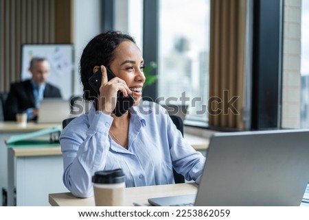 Young Latino businesswoman using phone while working in the office. Attractive young girl employee worker in formal wear sit on table, use computer and telephone feeling happy at corporate workplace. Royalty-Free Stock Photo #2253862059