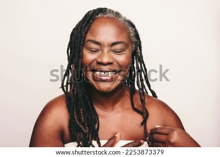 Cheerful mature woman laughing happily while wrapped in a bath towel. Refreshed woman with dreadlocks standing against a white background. Joyful middle aged woman taking care of her ageing body. Royalty-Free Stock Photo #2253873379