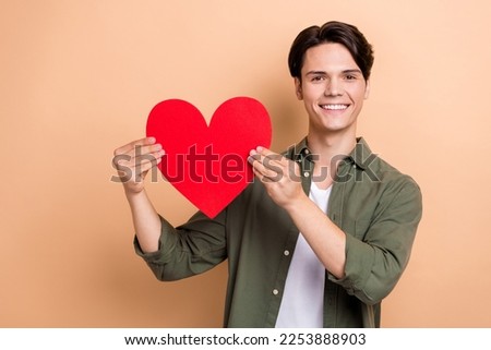 Photo of cheerful smiling man brunet hair wear khaki stylish shirt hold postcard gift valentine day holiday shape isolated on beige color background