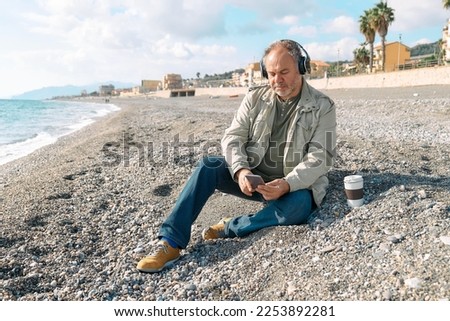 Happy middle-aged bearded man listening music or podcast from smartphone application in headphones while sitting on winter beach.