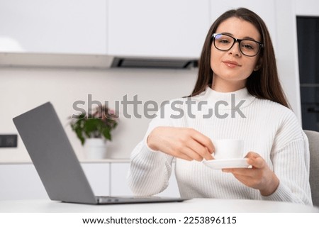 Portrait of a young beautiful woman holding a coffee cup and sitting in the kitchen near the laptop. Freelancer has a break 