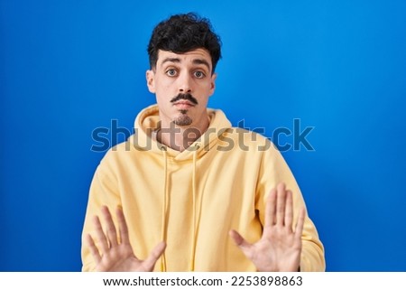 Hispanic man standing over blue background moving away hands palms showing refusal and denial with afraid and disgusting expression. stop and forbidden. 