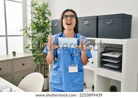Young hispanic doctor woman holding safety mask at clinic angry and mad screaming frustrated and furious, shouting with anger looking up. 