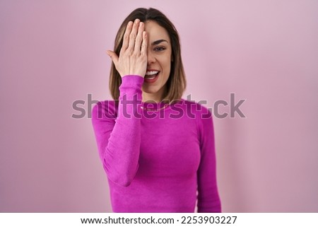 Hispanic woman standing over pink background covering one eye with hand, confident smile on face and surprise emotion. 
