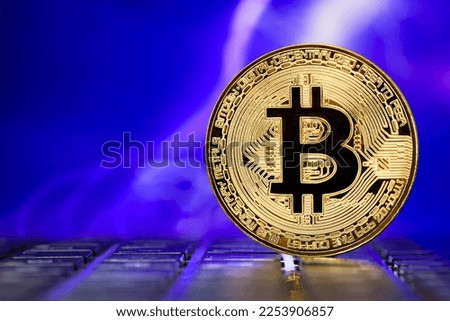 Golden metallic Bitcoin coin standing on a laptop keyboard against the background of a blurred dramatic multicolored stormy sky with lightning