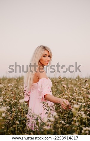 Vertical photo blonde woman posing in the big endless field of daisies in summer evening. Lady wear pink dressed, looking aside smiling, touching the plant green blossoming flower. Moody atmosphere
