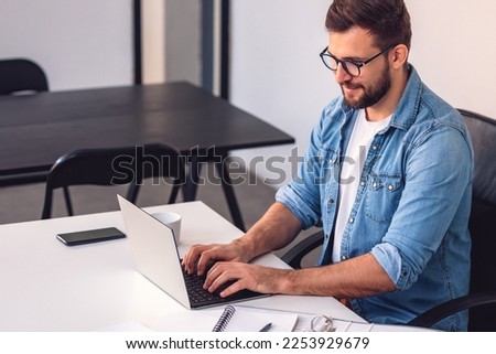 A smiling man with glasses is using a laptop in the office while sitting at his desk. Royalty-Free Stock Photo #2253929679