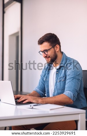 A smiling man with glasses is using a laptop in the office while sitting at his desk. Royalty-Free Stock Photo #2253929691