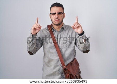 Young hispanic man wearing suitcase pointing up looking sad and upset, indicating direction with fingers, unhappy and depressed. 