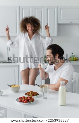 cheerful man with closed eyes drinking coffee near girlfriend dancing on blurred background