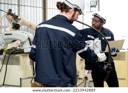 Industrial engineer checking robotic welder operation in modern automation factory. Diverse ethnic technician using laptop to maintenance, repair robot controller system for automated steel welding.
