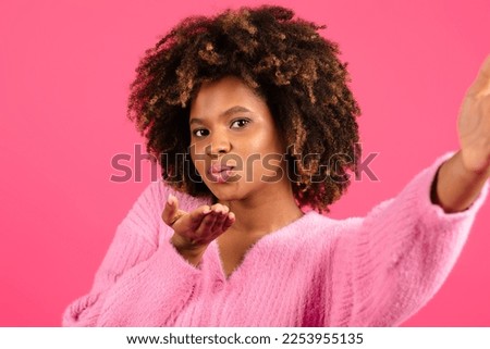 Cheerful young black curly lady in casual taking selfie, blowing kiss at camera isolated on pink background, close up, studio. Photo for blog and social networks, blogger shooting video, offer and ad