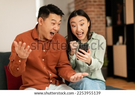 Portrait of overjoyed asian couple using smartphone and making winner sign gesture, shaking fists, sitting on couch at home, reading great news Royalty-Free Stock Photo #2253955139