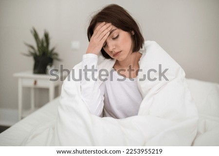 Sick unhappy young beautiful brunette woman waking up with headache in the morning, ill lady sitting in bed covered in blanket at home, touching her head, suffering from flu or coronavirus, copy space Royalty-Free Stock Photo #2253955219